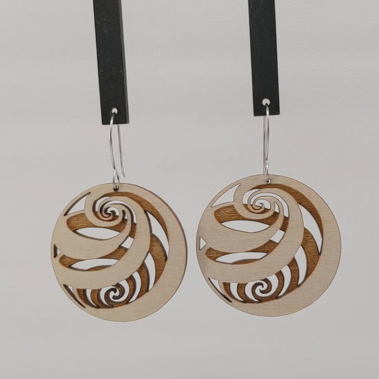 Pair of wooden earrings with silver stainless steel hooks. They are light finish balls with spirals engraved creating a 3D spherical illusion. Made from birch wood hanging and rotating against a white background.
