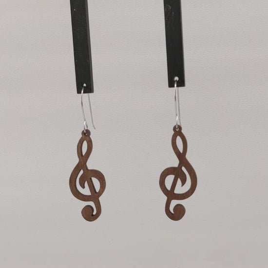 Pair of wooden earrings with silver stainless steel hooks. They are brown cutouts of the musical notes treble clef. Made from birch wood hanging and rotating against a white background.