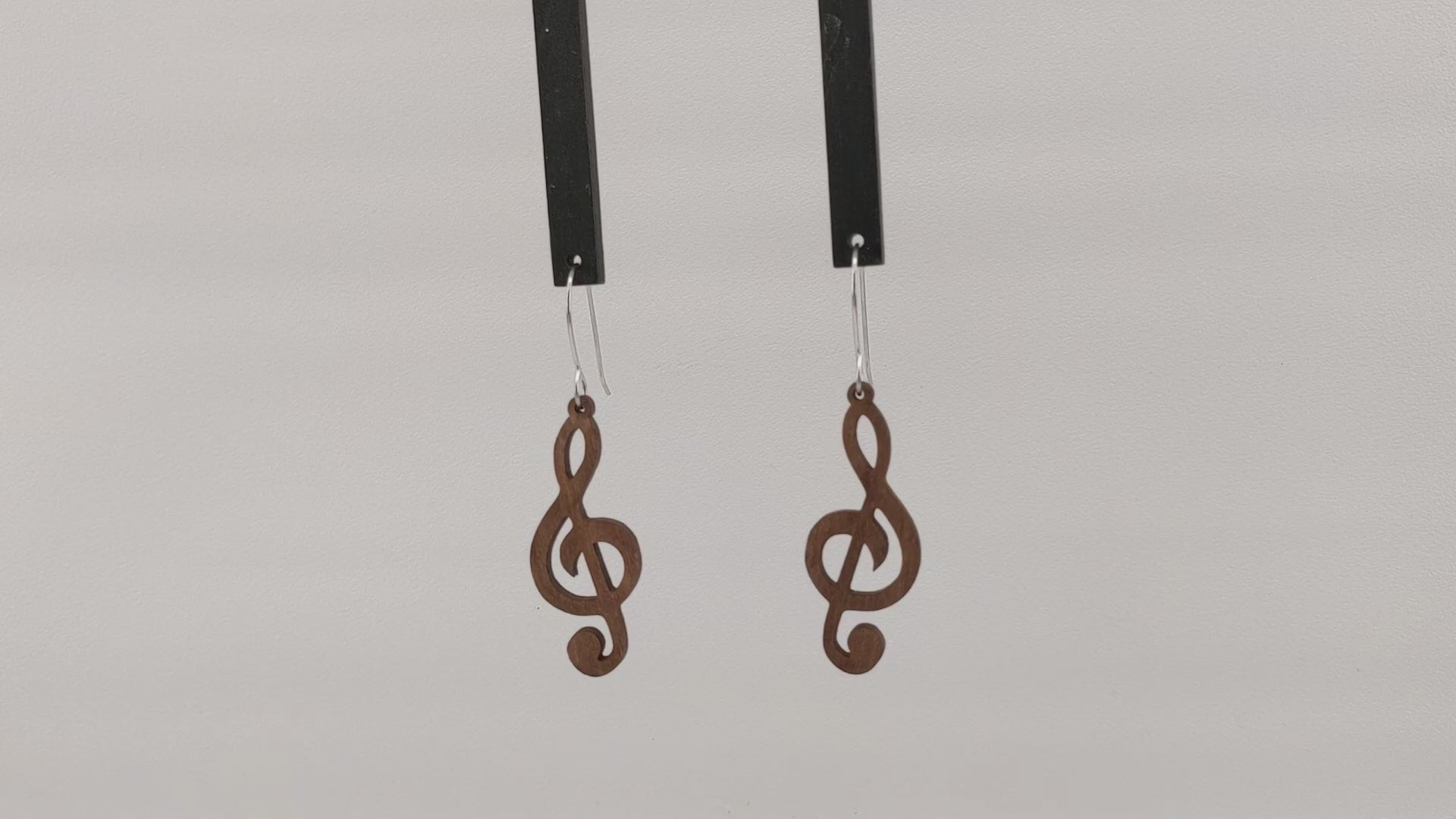 Pair of wooden earrings with silver stainless steel hooks. They are brown cutouts of the musical notes treble clef. Made from birch wood hanging and rotating against a white background.