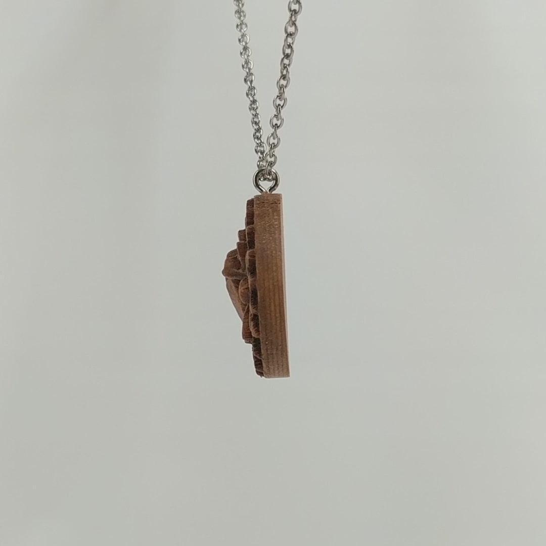 Wooden necklace pendant carved in the shape of a honey bee resting on a flower. Made from hard maple wood and hanging from a silver stainless steel chain against a white background rotating on a table.