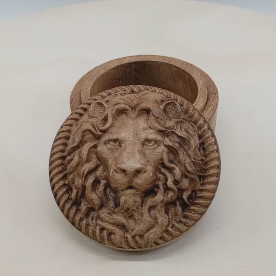 Round wooden box 3D carved with a lions face looking straight ahead surrounded by a rope border. With stern look in its eyes with its mane parted in the middle draping over its face. Made from hard maple wood stained brown against a white background rotating on a table.
