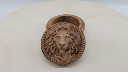 Round wooden box 3D carved with a lions face looking straight ahead surrounded by a rope border. With stern look in its eyes with its mane parted in the middle draping over its face. Made from hard maple wood stained brown against a white background rotating on a table.