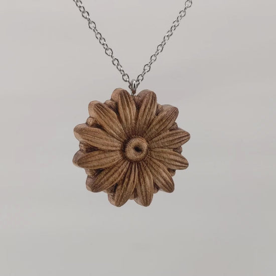 Wooden necklace pendant carved into the shape of a daisy flower.  Hanging and rotating from a silver stainless steel chain against a white background.