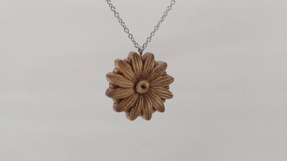 Wooden necklace pendant carved into the shape of a daisy flower.  Hanging and rotating from a silver stainless steel chain against a white background.