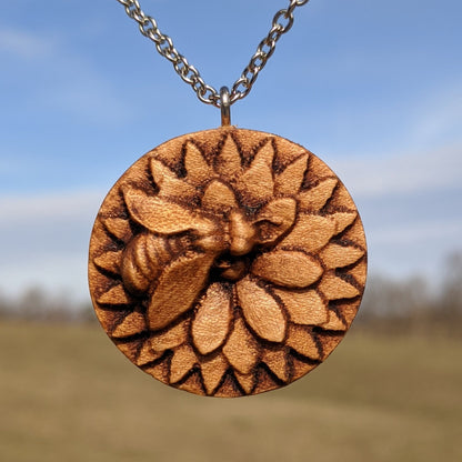 Wooden necklace pendant carved in the shape of a honey bee resting on a flower. Made from hard maple wood and hanging from a silver stainless steel chain against a landscape background background.