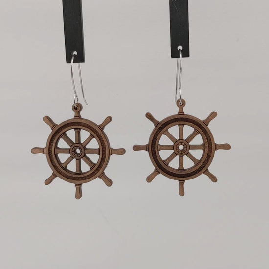 Pair of wooden earrings with silver stainless steel hooks. They are brown pirate ship wheels with 8 handles and a spoked center. Made from birch wood hanging and rotating against a white background.