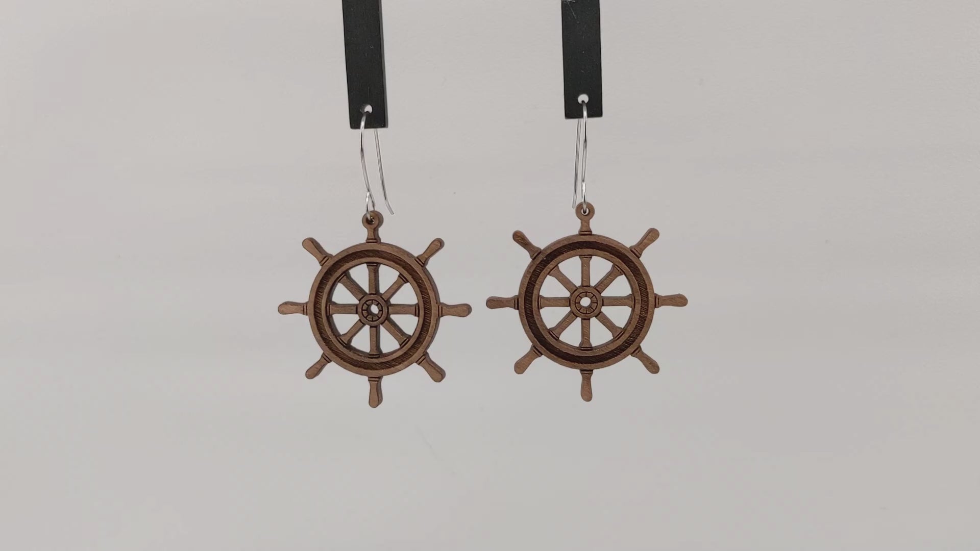 Pair of wooden earrings with silver stainless steel hooks. They are brown pirate ship wheels with 8 handles and a spoked center. Made from birch wood hanging and rotating against a white background.