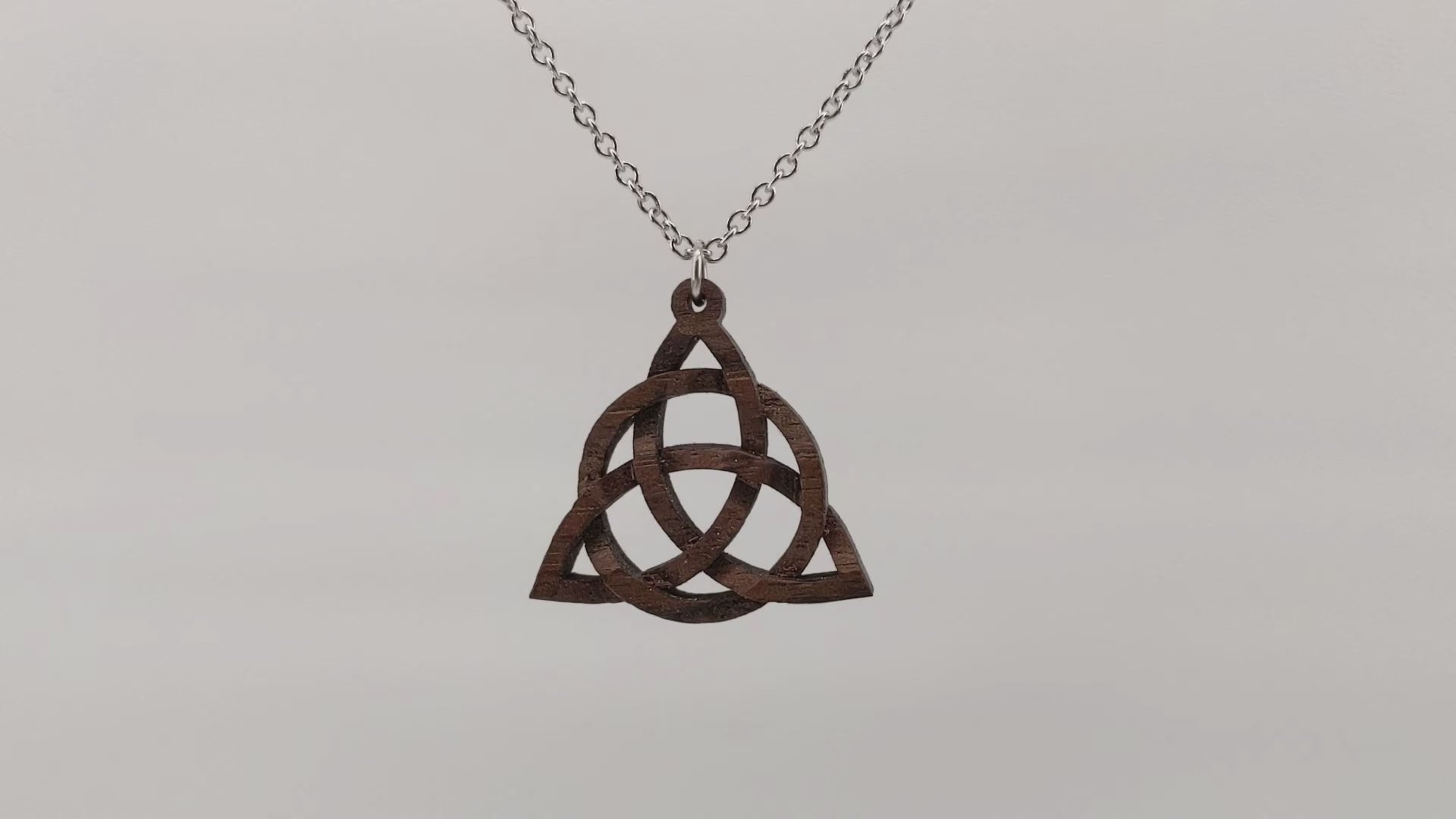 Wooden necklace pendant created in the style of a Celtic woven trinity symbol. Made from hard maple. Hanging and rotating from a silver stainless steel chain against a white background.