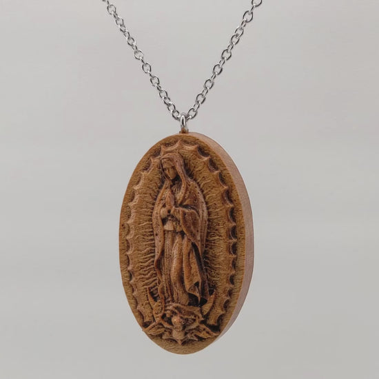 Wooden necklace pendant carved in the shape of Guadalupe. Made from hard maple wood. Hanging and rotating from a silver stainless steel chain against a white background.