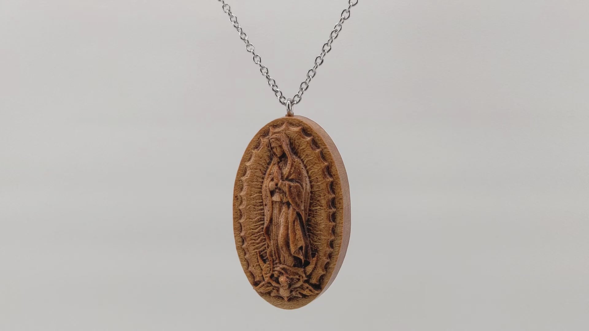 Wooden necklace pendant carved in the shape of Guadalupe. Made from hard maple wood. Hanging and rotating from a silver stainless steel chain against a white background.