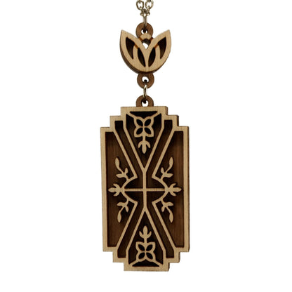 Wooden necklace pendant made from two contrasting layers of birch wood. The top lighter layer is intricately cut with flower like details. The overall shape is rectangular. It hangs from a silver stainless steel chain against a white background.