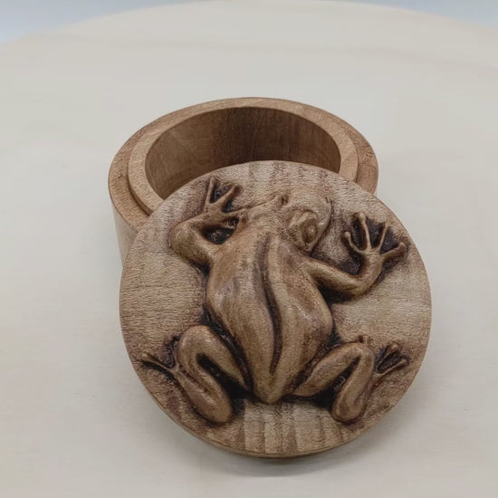 Round wooden box 3D carved with a tree frog in a clinging position. It has smooth skin and long skinny limbs and hands with bulbous fingertips Made from hard maple wood with a brown stain against a white background rotating on a table.