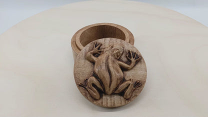 Round wooden box 3D carved with a tree frog in a clinging position. It has smooth skin and long skinny limbs and hands with bulbous fingertips Made from hard maple wood with a brown stain against a white background rotating on a table.