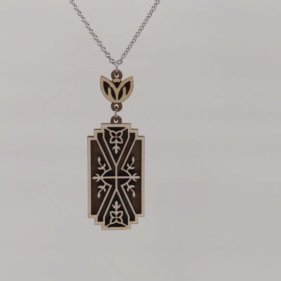 Wooden necklace pendant made from two contrasting layers of birch wood. The top lighter layer is intricately cut with flower like details. The overall shape is rectangular. Hanging and rotating from a silver stainless steel chain against a white background.