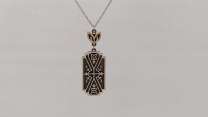 Wooden necklace pendant made from two contrasting layers of birch wood. The top lighter layer is intricately cut with flower like details. The overall shape is rectangular. Hanging and rotating from a silver stainless steel chain against a white background.