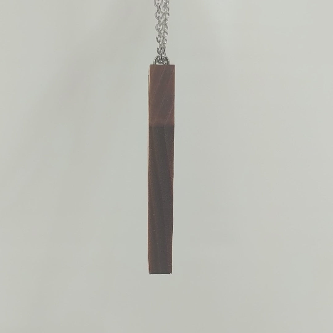 Wooden necklace pendant created in the shape a skeleton lying in a coffin. Hanging from a silver stainless steel chain against a white background rotating on a table.