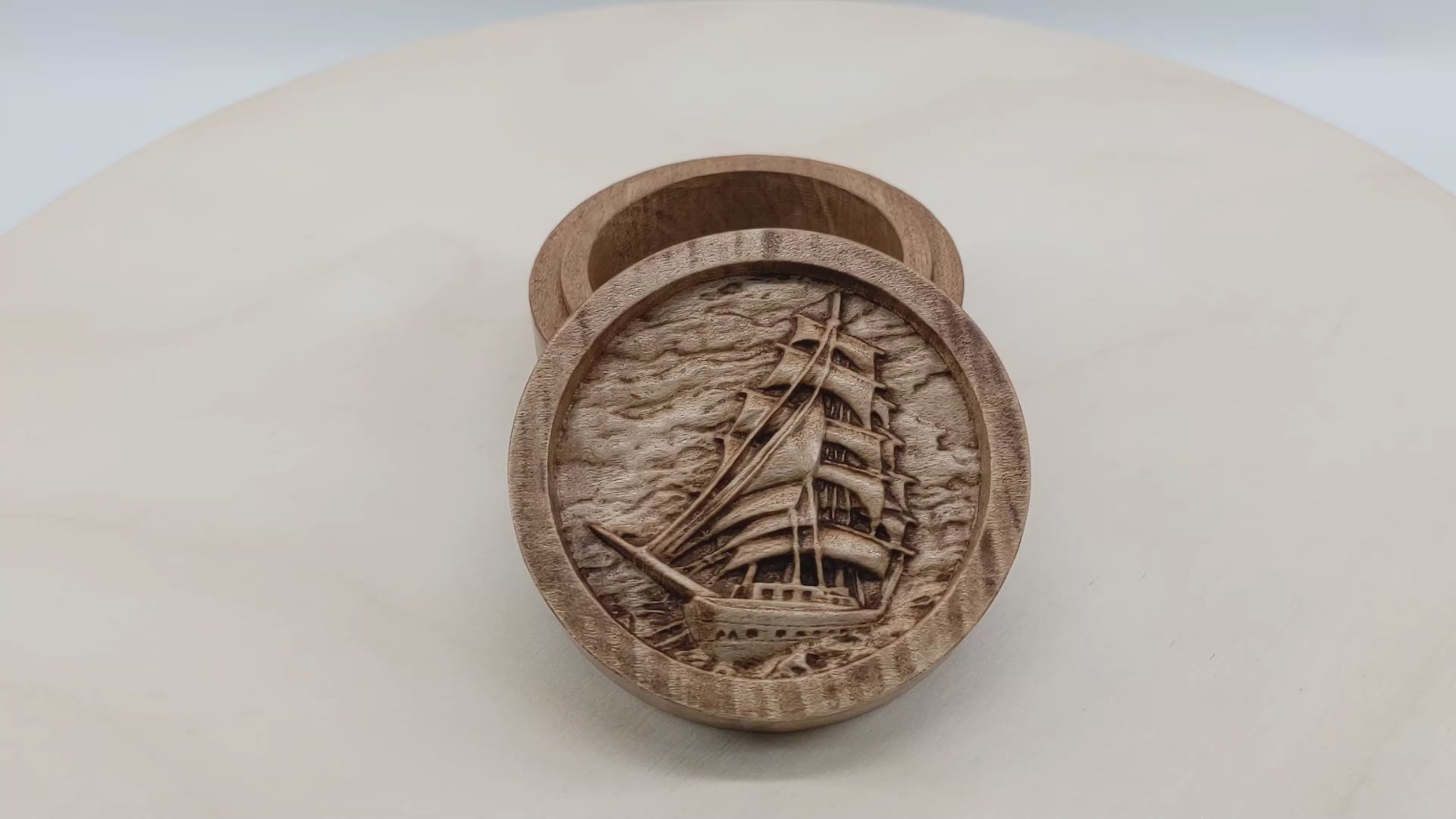 Round wooden box 3D carved with a large sailboat on the sea. It has multiple sails all pulled taught with ropes hanging all around. The background is a cloudy sky and rolling waves. Made from hard maple wood stained brown against a white background rotating on a table.