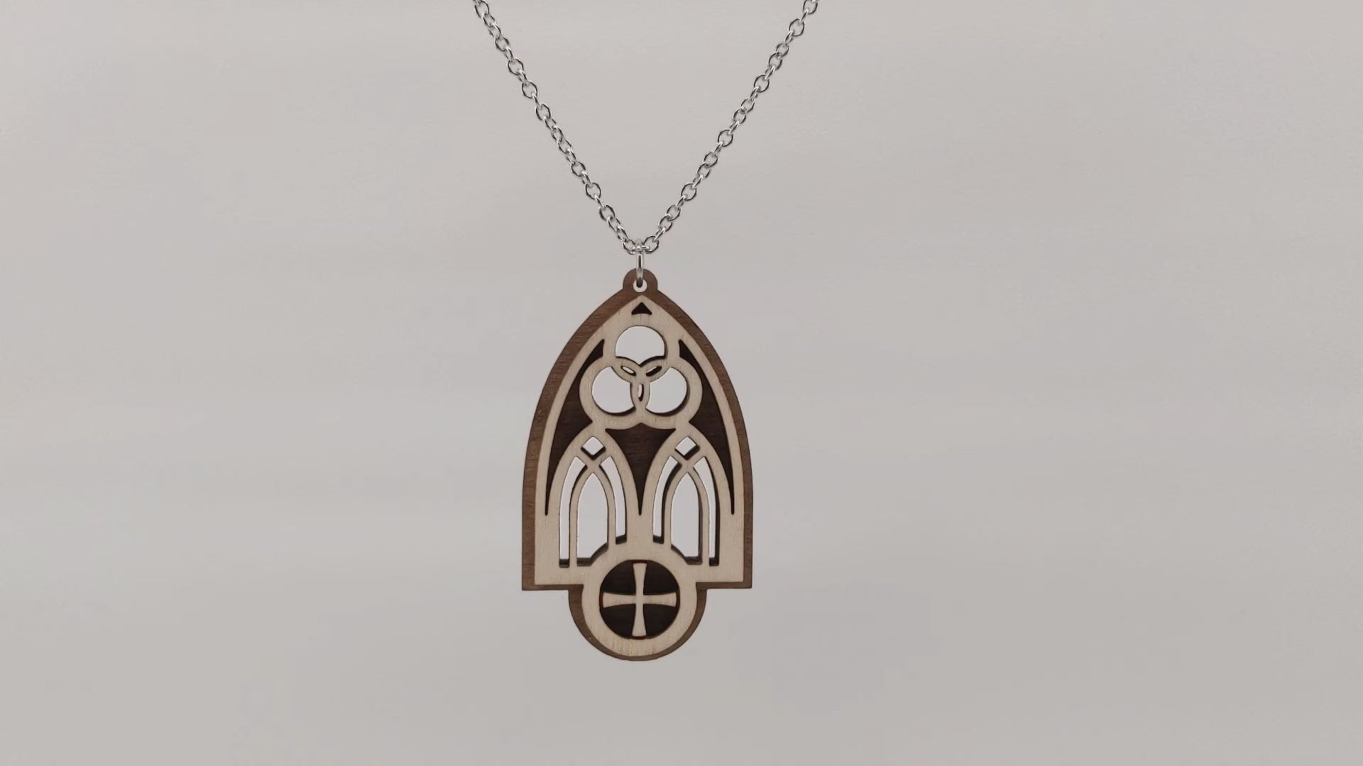 Wooden necklace pendant inspired by a gothic church window. includes a trinity symbol above and a cross below. Made from two layers, dark and light birch wood. Hanging and rotating from a silver stainless steel chain against a white background.