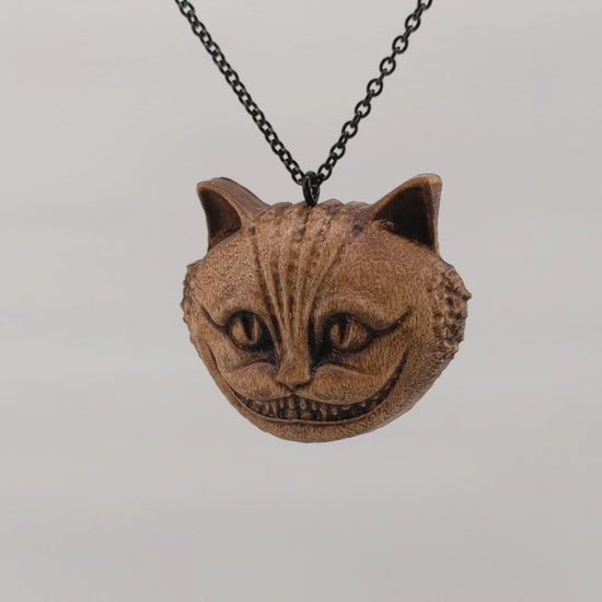 Wooden necklace pendant carved in the shape of a Cheshire cat head.  Hanging and rotating from a black stainless steel chain against a white background.