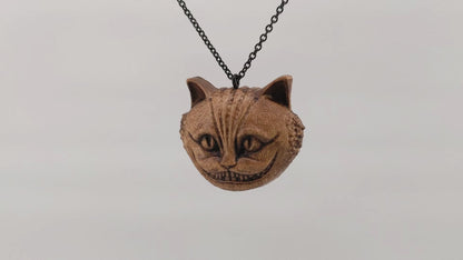 Wooden necklace pendant carved in the shape of a Cheshire cat head.  Hanging and rotating from a black stainless steel chain against a white background.