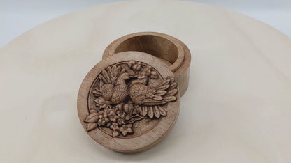 Round wooden box 3D carved with two lovebirds standing on a branch blooming with flowers, looking at each other with their beaks touching and feathers fluffed up. Made from hard maple wood stained brown against a white background rotating on a table.