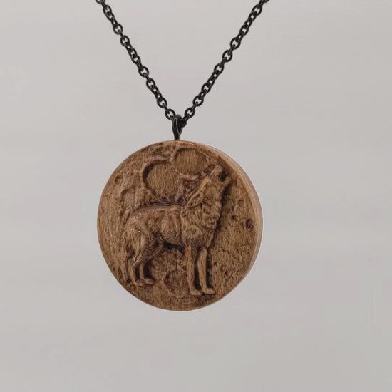 Wooden necklace pendant depicting a howling wolf in front of a full moon. Carved in a realistic style from hard maple wood. Hanging and rotating from a black stainless steel chain against a white background.