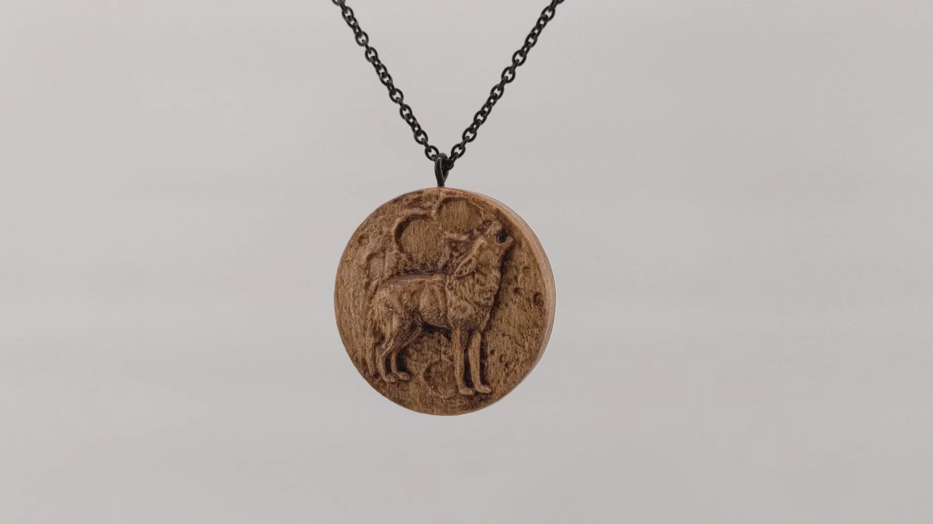 Wooden necklace pendant depicting a howling wolf in front of a full moon. Carved in a realistic style from hard maple wood. Hanging and rotating from a black stainless steel chain against a white background.