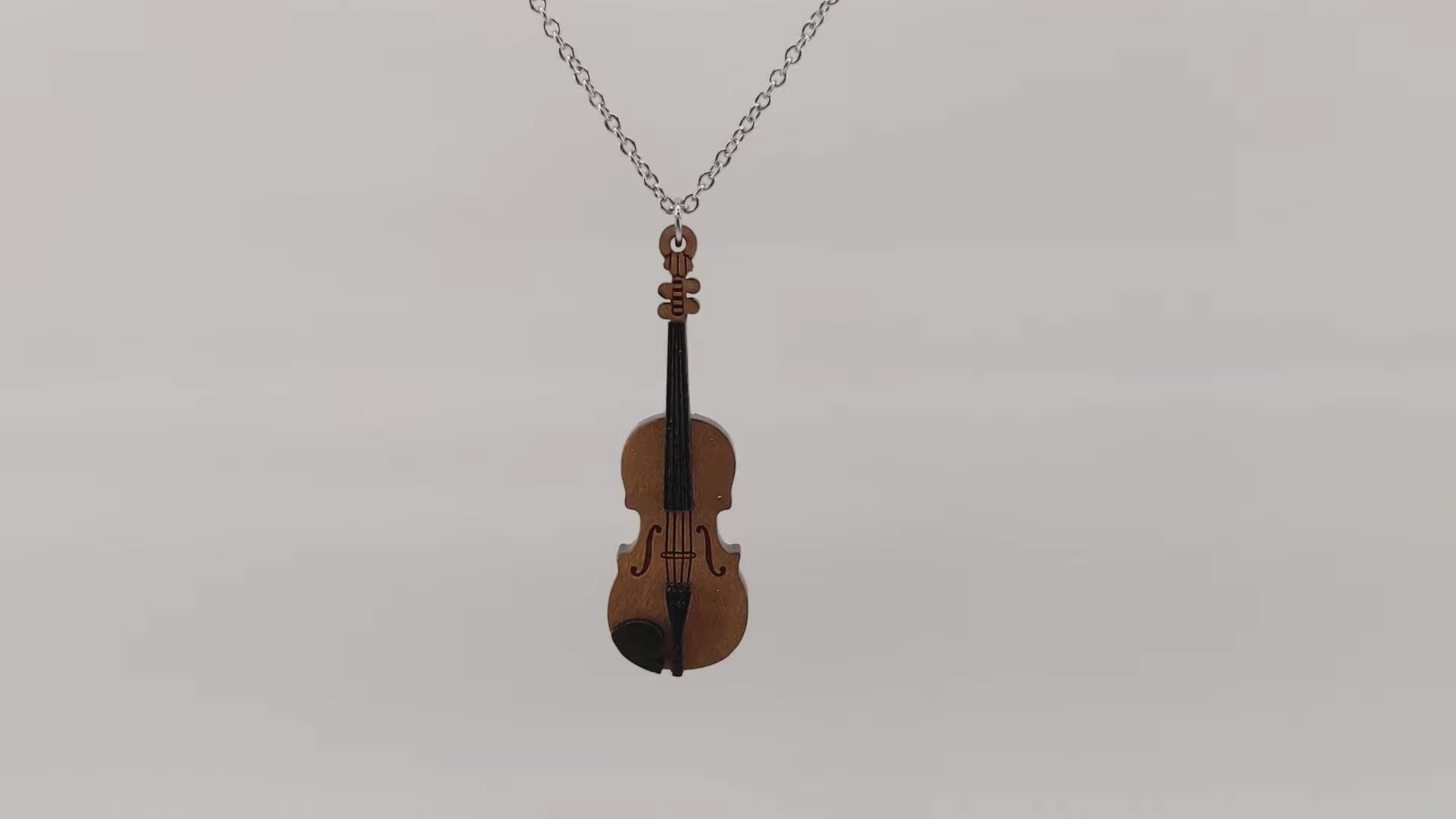 Wooden necklace pendant created in the shape of a miniature violin. Made from hard maple wood. Hanging and rotating from a silver stainless steel chain against a white background.