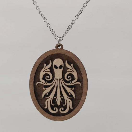 Oval wooden octopus pendant necklace made of contrasting layers of light and dark birch wood. The Octopus is light in color overlayed onto a darker base. Hanging and rotating from a silver stainless steel chain against a white background.