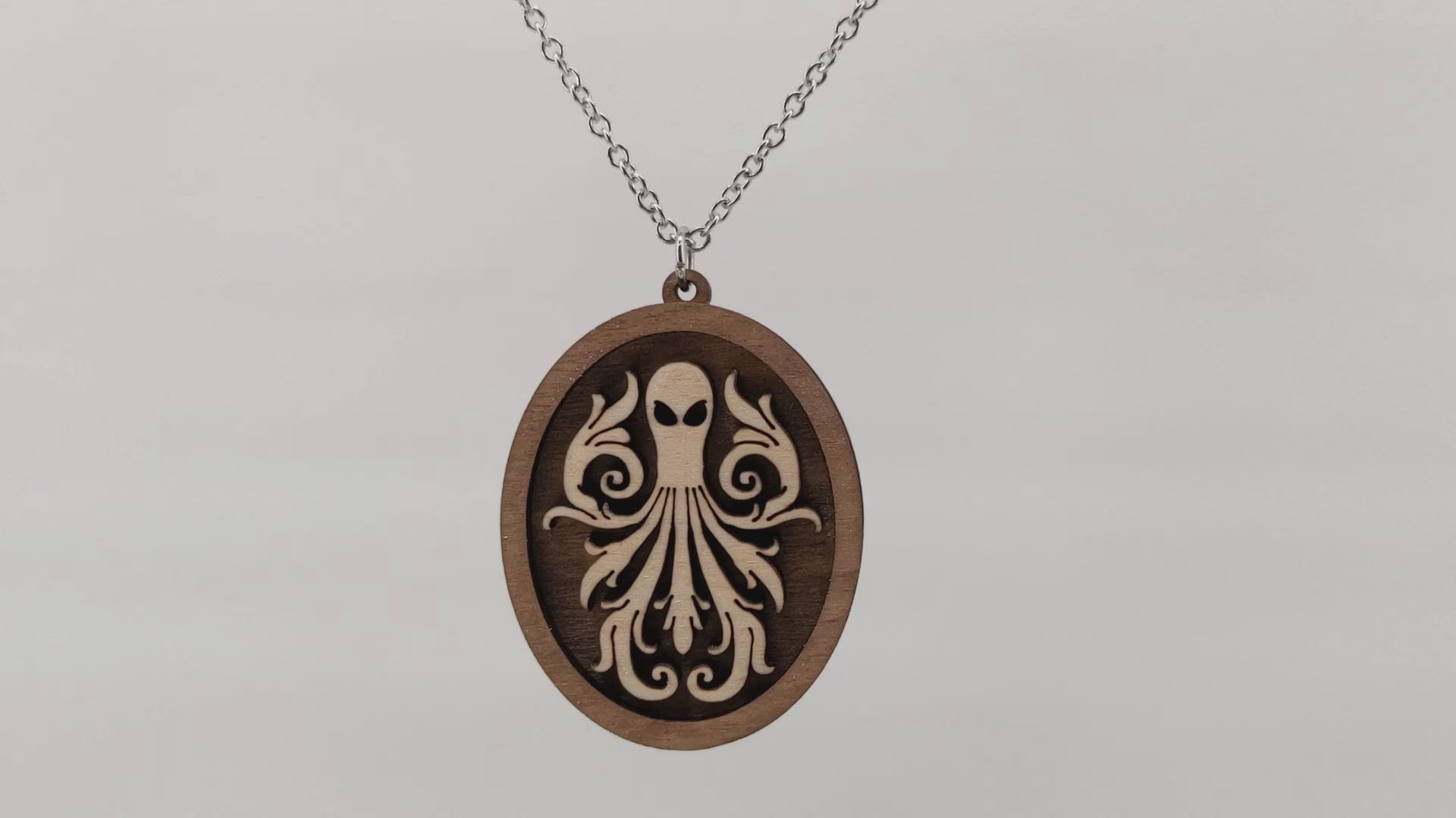 Oval wooden octopus pendant necklace made of contrasting layers of light and dark birch wood. The Octopus is light in color overlayed onto a darker base. Hanging and rotating from a silver stainless steel chain against a white background.