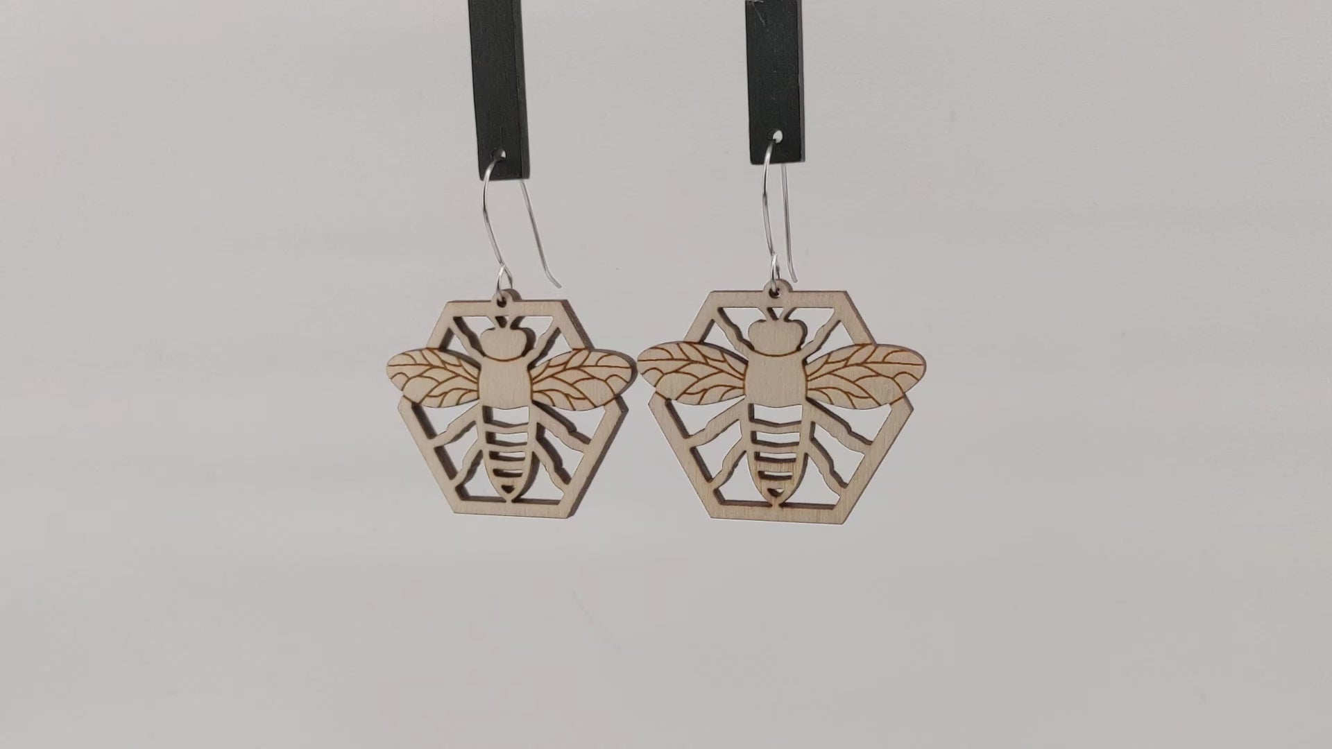 Pair of wooden earrings with silver stainless steel hooks. They are natural finished honeybees with its arms and wings spread on a honeycomb. Made from birch wood hanging and rotating against a white background.