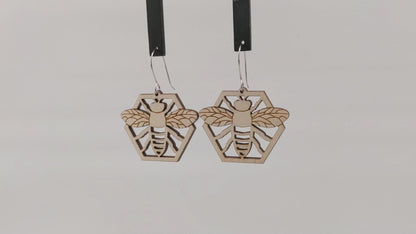 Pair of wooden earrings with silver stainless steel hooks. They are natural finished honeybees with its arms and wings spread on a honeycomb. Made from birch wood hanging and rotating against a white background.