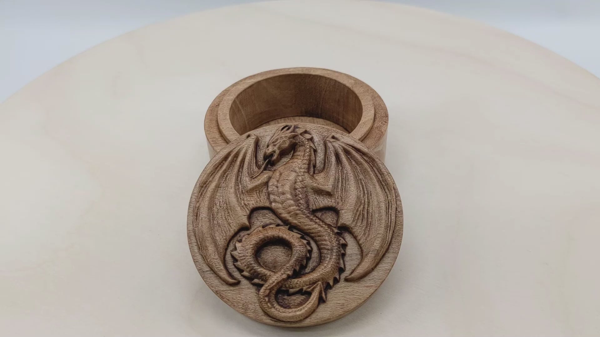 Round wooden box 3D carved with a mediaeval dragon with pointed wings spread, horned head, open mouth and serpentine tongue. It has scales down its belly and spines all down its back. Made from hard maple wood stained brown against a white background rotating on a table.