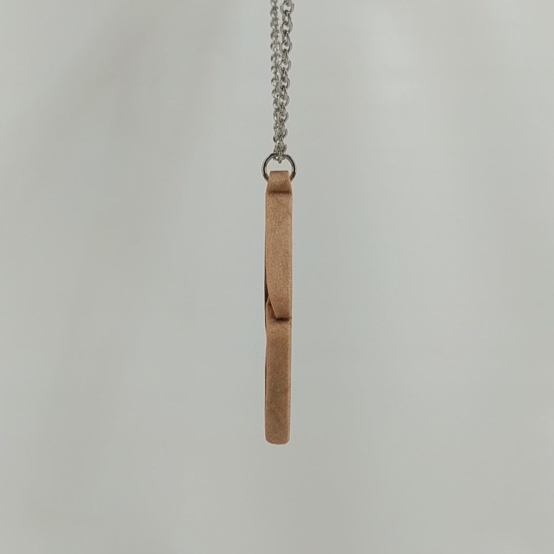 Wooden necklace pendant carved so that curving shapes weave into each other. Made from hard maple wood and hanging from a silver chain against a white background rotating on a table. 