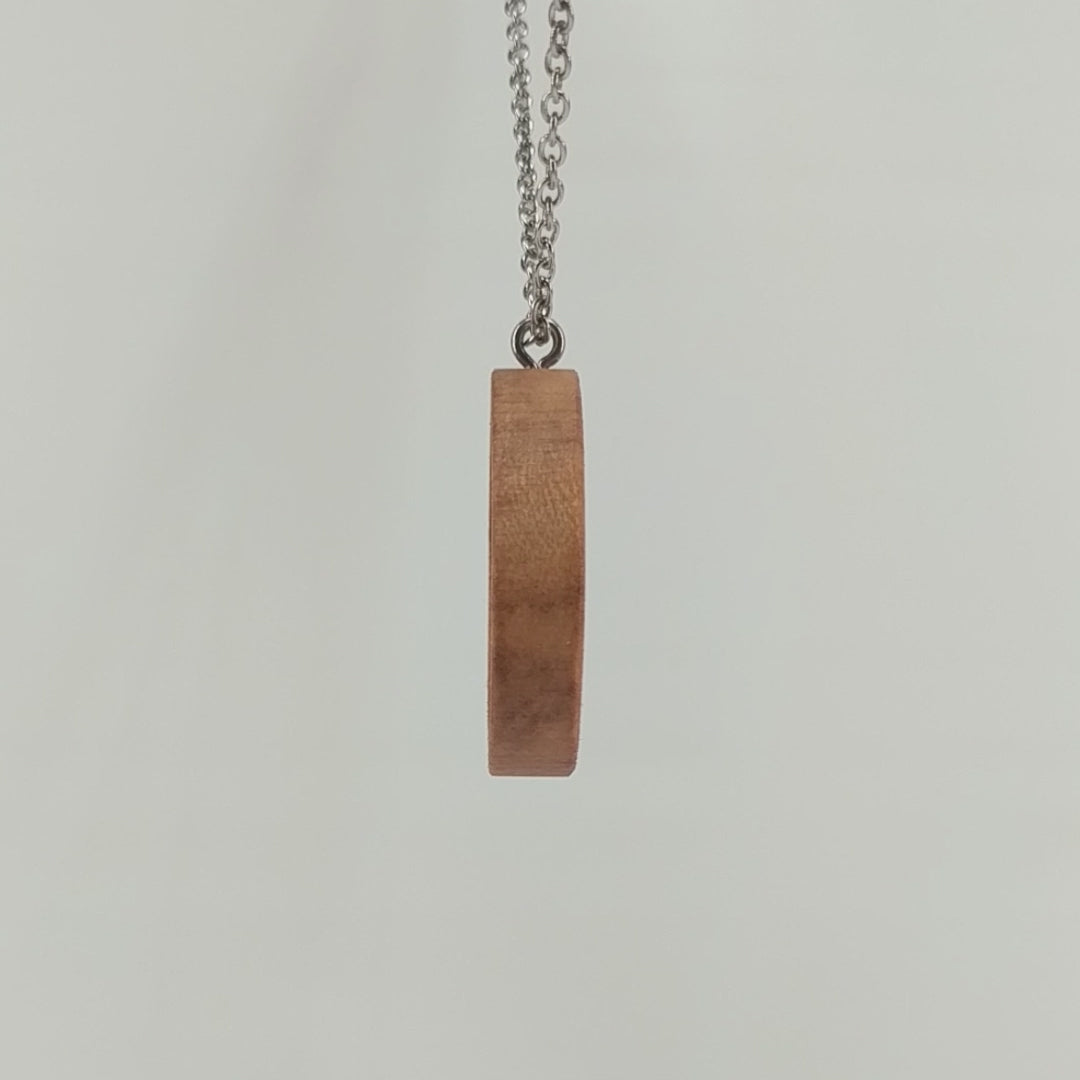 Round wooden necklace pendant carved in the shape of two hands reaching out to touch each other. Made from hard maple wood. Hanging from a silver stainless steel chain against a white background rotating on a table.