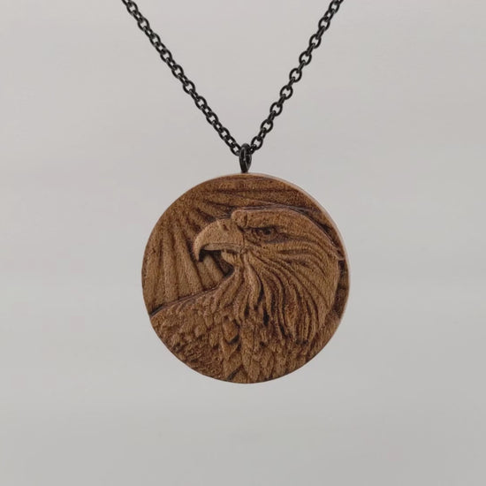 Round wooden necklace pendant carved in the shape of an eagles head. Made from hard maple wood. Hanging and rotating from a black stainless steel chain against a white background.