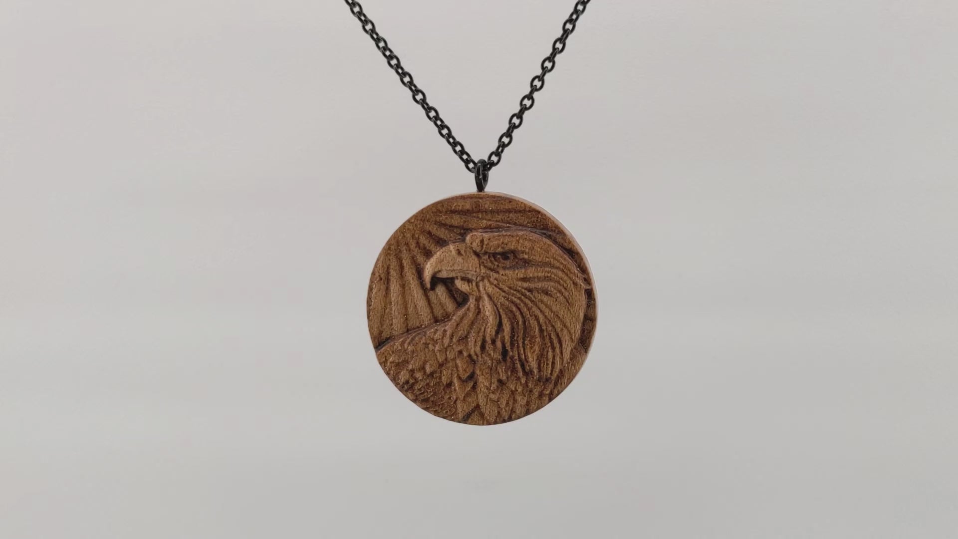 Round wooden necklace pendant carved in the shape of an eagles head. Made from hard maple wood. Hanging and rotating from a black stainless steel chain against a white background.