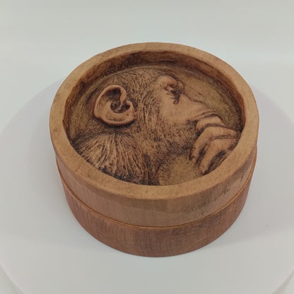 Round wooden box 3D carved with a recessed carving of a side profile view of an hairy ape rubbing his chin in his hand while in mid thought. Made from hard maple wood with a brown stain against a white background rotating on a table.