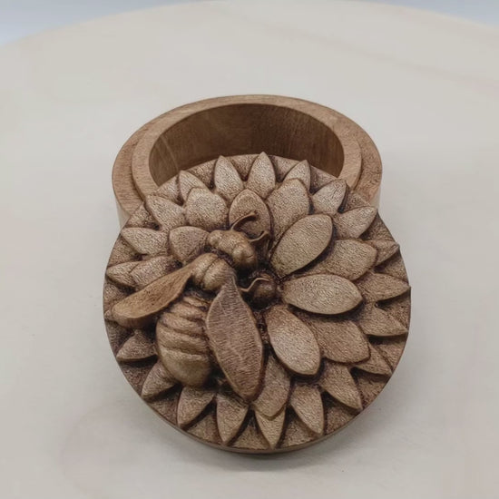 Round wooden box 3D carved with a honey bee rested atop a multi-layered flower. The bee is curved slightly off from the center of the flower as if searching for nectar. Made from hard maple wood with a brown stain against a white background rotating on a table.