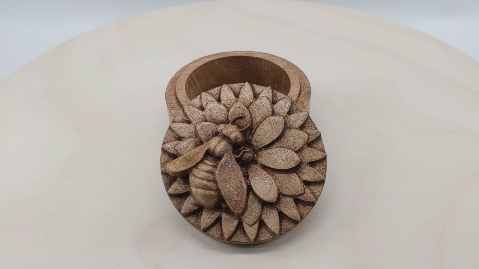 Round wooden box 3D carved with a honey bee rested atop a multi-layered flower. The bee is curved slightly off from the center of the flower as if searching for nectar. Made from hard maple wood with a brown stain against a white background rotating on a table.