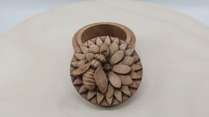 Round wooden box 3D carved with a honey bee rested atop a multi-layered flower. The bee is curved slightly off from the center of the flower as if searching for nectar. Made from hard maple wood with a brown stain against a white background rotating on a table.