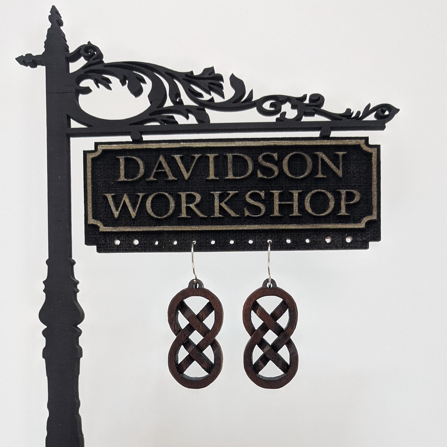 Pair of wooden earrings with silver stainless steel hooks. They are Celtic style, carved in a woven pattern making a figure 8 with exes inside. Made from dark walnut wood hanging from a model Davidson Workshop sign against a white background.