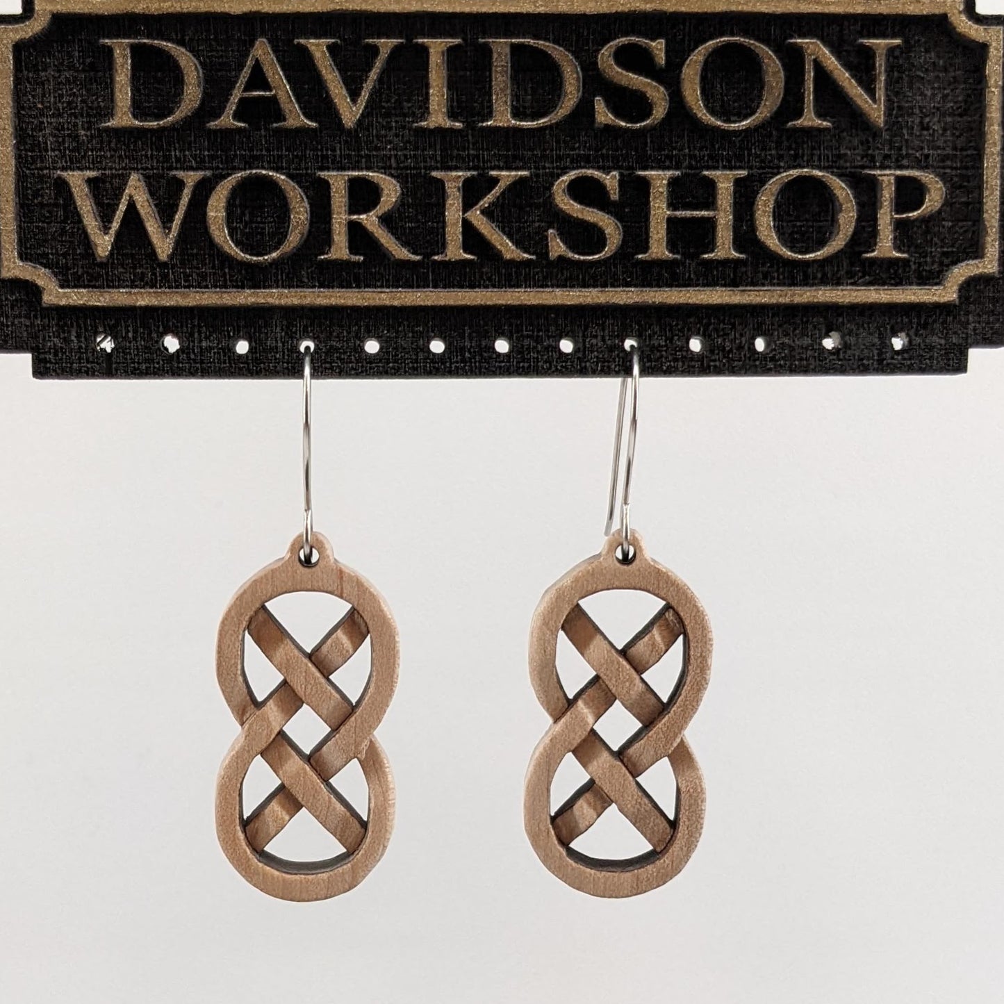 Pair of wooden earrings with silver stainless steel hooks.They are Celtic style, carved in a woven pattern making a figure 8 with exes inside. Made from hard maple hanging from a model Davidson Workshop sign against a white background (Close up View)