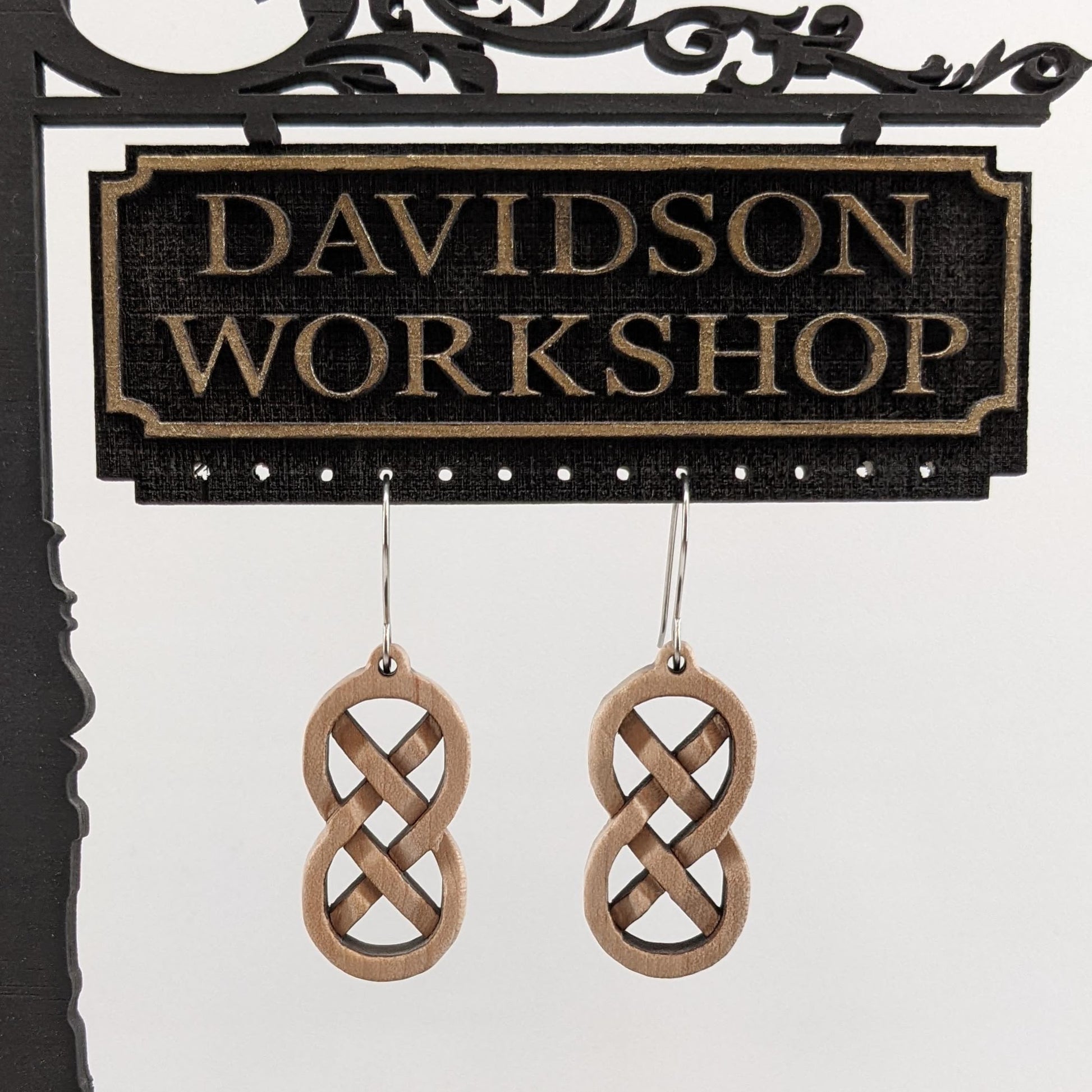 Pair of wooden earrings with silver stainless steel hooks.They are Celtic style, carved in a woven pattern making a figure 8 with exes inside. Made from hard maple hanging from a model Davidson Workshop sign against a white background.