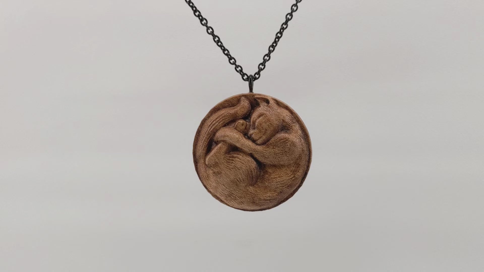 Wooden necklace pendant carved into the shape of a cat curled into a round ball. Hanging and rotating from a black stainless steel chain against a white background.