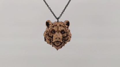 Wooden necklace pendant carved in the shape of a grizzly bears head. Made from hard maple wood. Hanging and rotating from a black stainless steel chain against a white background.