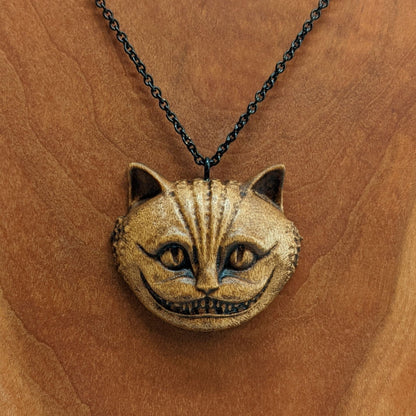 Wooden necklace pendant carved in the shape of a Cheshire cat head. Hanging from a black stainless steel chain against a cherry wood background.