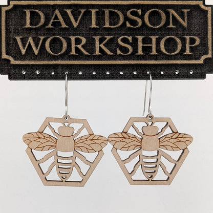 Pair of wooden earrings with silver stainless steel hooks. They are natural finished honeybees with its arms and wings spread on a honeycomb. Made from birch wood hanging from a model Davidson Workshop sign against a white background (Close up View)