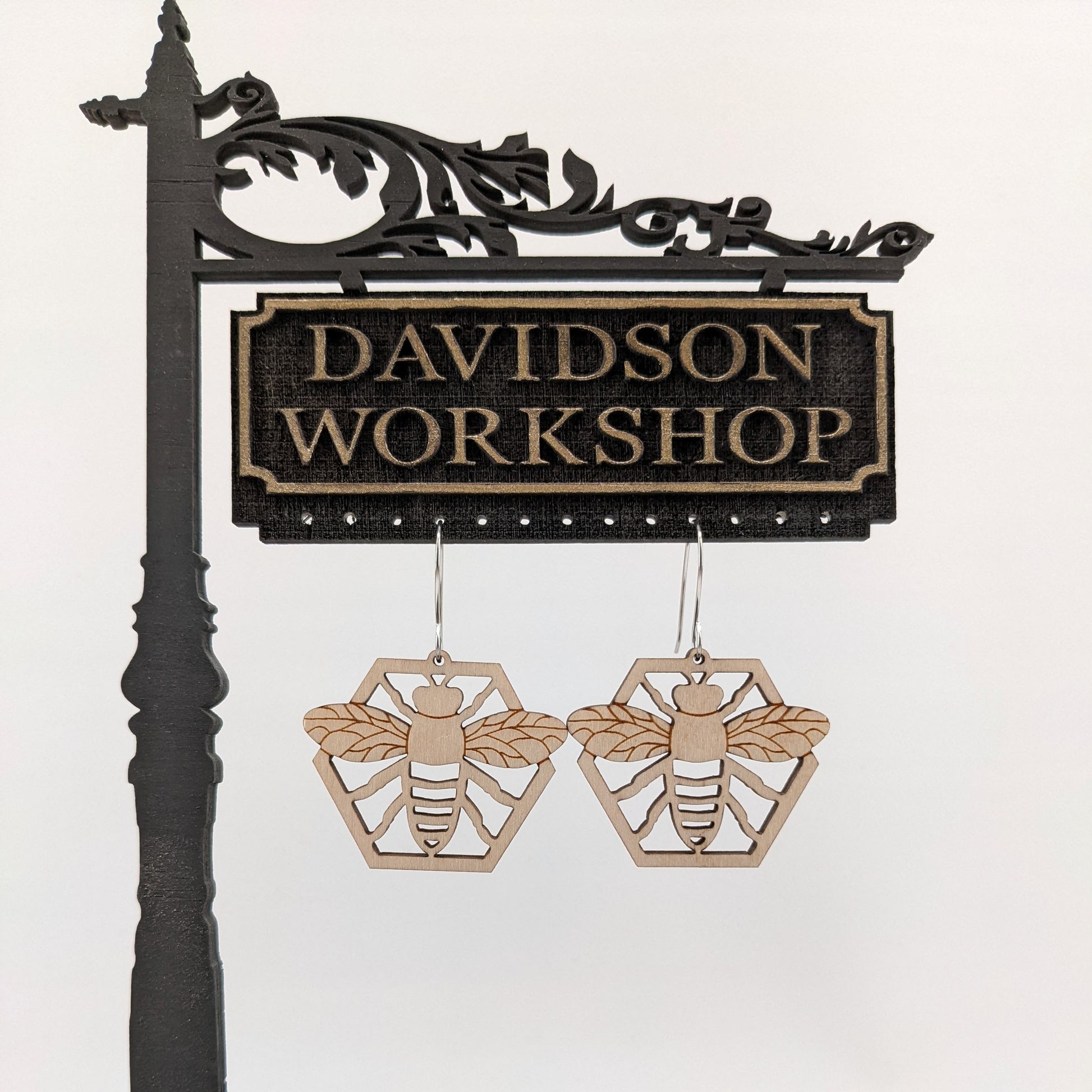 Pair of wooden earrings with silver stainless steel hooks. They are natural finished honeybees with its arms and wings spread on a honeycomb. Made from birch wood hanging from a model Davidson Workshop sign against a white background.