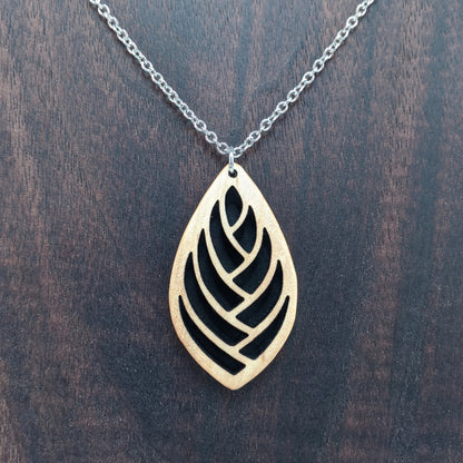 Wooden necklace pendant cut into a teardrop shape with a herring bone pattern cut throughout it. Made from hard maple and hanging from a silver stainless steel chain against a cherry wood background.